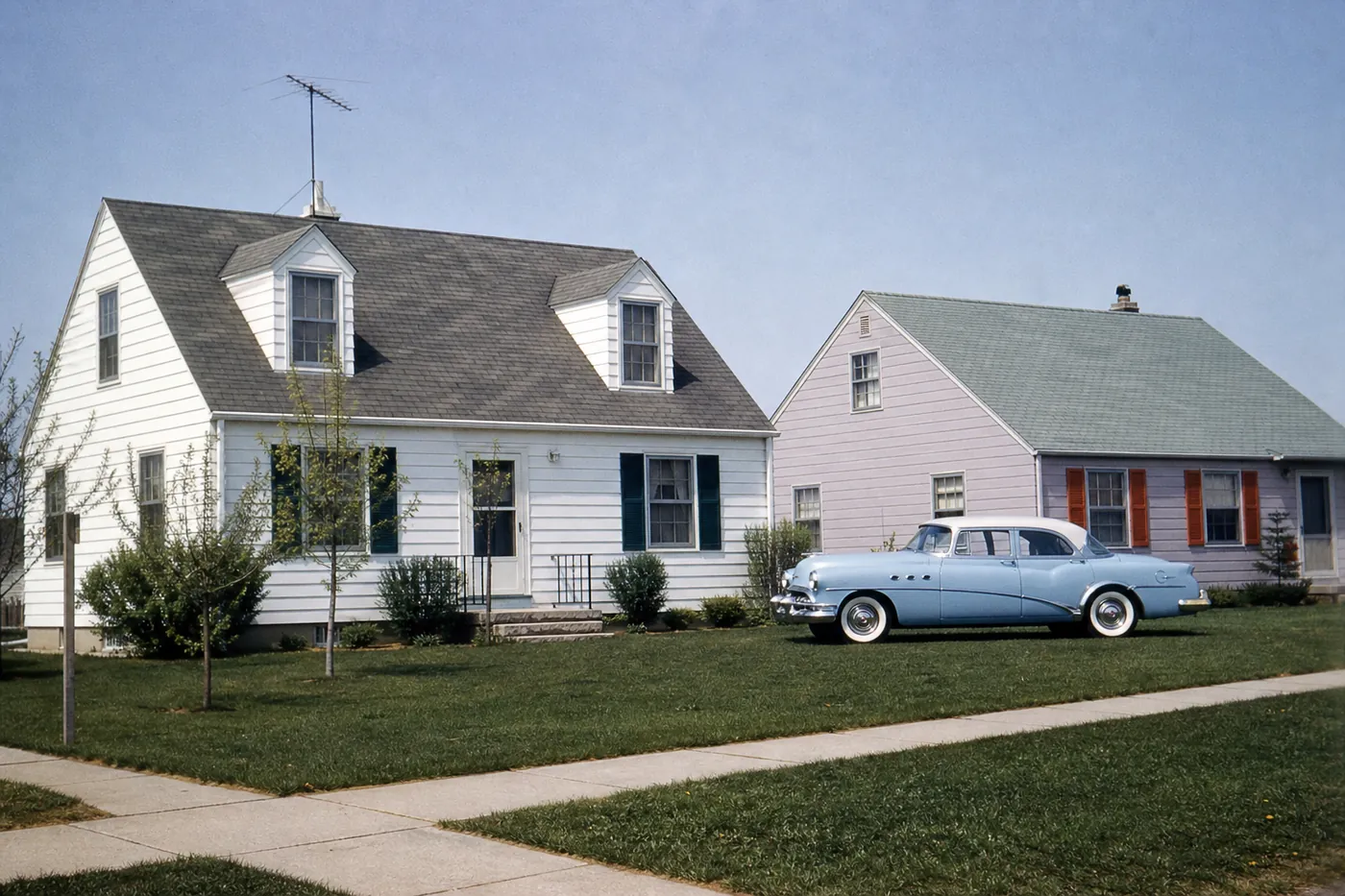Kodachrome-style still of two mid-century houses and a pale blue car on a sunny street.
